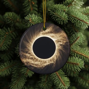 Puede incluir: Un adorno redondo de cerámica con una imagen negra y dorada de un eclipse solar. El eclipse está rodeado por un halo dorado brillante.