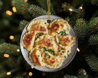 Puede incluir: Adorno navideño redondo con un plato de pierogi, una especialidad polaca. Las empanadillas son de color marrón dorado con un relleno y espolvoreadas con hierbas verdes. El adorno cuelga de un hilo dorado sobre un árbol de Navidad.