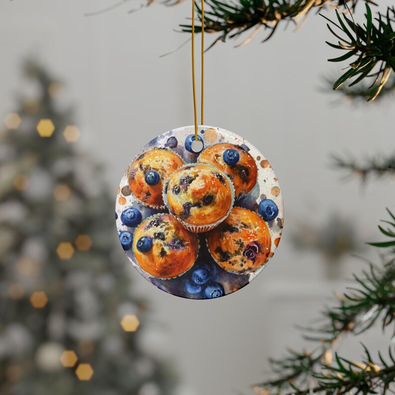Baking blueberry muffins round ceramic ornament featuring a detailed image of freshly baked blueberry muffins with golden tops and scattered berries. The glossy finish enhances the warm tones of the muffins. High quality ceramic ornament hanging by a gold string. Pine tree in the background with lights behind the ornament.