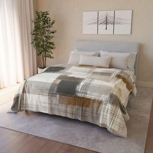 May include: A bed with a patterned blanket in neutral tones of gray, brown, and cream. The blanket features a brushstroke design. The bed is made up with white pillows and a gray headboard. A tree in a pot is to the left of the bed.