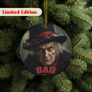 May include: A round Christmas ornament featuring a stern-faced man in a black hat and coat, with a red scarf. The word "BAH" is in red block letters. The ornament is hanging from a gold string and has the words "Limited Edition" in red.