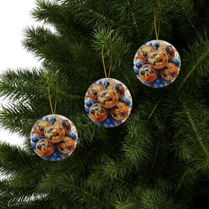 Baking blueberry muffins round ceramic ornaments featuring three glossy ceramic pieces each showing detailed images of golden blueberry muffins with scattered berries. All three ornaments hang neatly by gold strings on the same branch. Pine tree in the background behind the ornaments.
