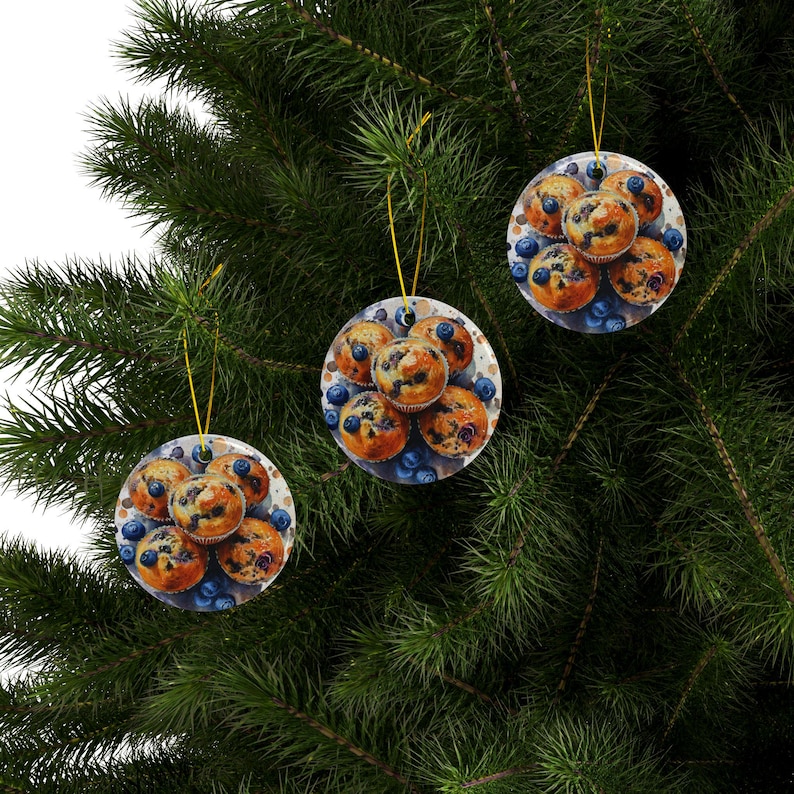 Baking blueberry muffins round ceramic ornaments featuring three glossy ceramic pieces each showing detailed images of golden blueberry muffins with scattered berries. All three ornaments hang neatly by gold strings on the same branch. Pine tree in the background behind the ornaments.