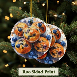 Baking blueberry muffins round ceramic ornament featuring a detailed image of freshly baked blueberry muffins with golden tops and scattered berries. The glossy finish enhances the warm tones of the muffins. High quality ceramic ornament hanging by a gold string. Pine tree in the background with lights behind the ornament. Showing the front and the back have the same print.