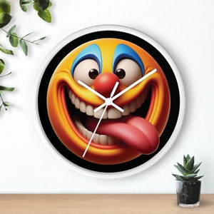 May include: A white clock with a black background and a yellow smiley face with a red nose and blue eyes. The smiley face is sticking its tongue out.