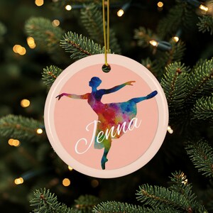 Op de afbeelding: Ronde kerstversiering met een ballerina in een kleurrijk aquarelontwerp. De versiering heeft een lichtroze achtergrond en de naam "Jenna" in witte letters. Een gouden draad is bevestigd om op te hangen.