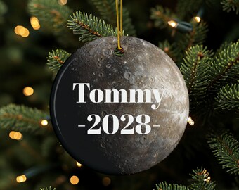 May include: A round Christmas ornament with a black border and a detailed image of a planet. The text "Tommy -2028-" is in white. The ornament hangs from a gold string against a backdrop of a Christmas tree.