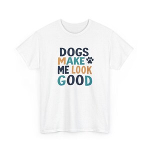 May include: A white short-sleeved t-shirt featuring the text DOGS MAKE ME LOOK GOOD in dark blue, tan, and teal lettering, accompanied by a small dark blue paw print icon positioned to the right of the word MAKE.
