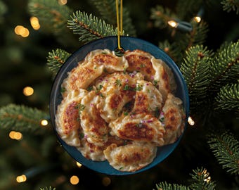 Puede incluir: Adorno navideño redondo que representa un cuenco azul lleno de pierogi. Los pierogi son de color marrón dorado y están cubiertos con cebollas rojas y hierbas. El adorno cuelga de un hilo dorado sobre un fondo de árbol de Navidad.