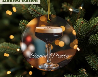 Può includere: Ornamento natalizio rotondo con un cocktail e la scritta "Sippin Pretty". L'ornamento ha un cordino dorato per appenderlo e la scritta "Limited Edition" in alto. Lo sfondo è sfocato con luci e rami di pino.
