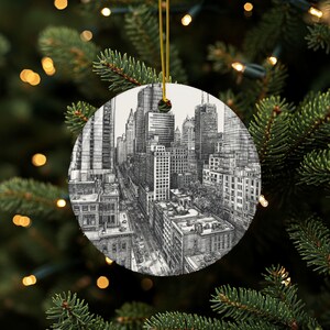 Puede incluir: Adorno navideño redondo y blanco con un dibujo detallado en blanco y negro de un horizonte urbano. El adorno está suspendido por un hilo dorado y se encuentra sobre un fondo de un árbol de Navidad con luces.
