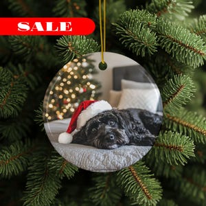 Könnte beinhalten: Runder Weihnachtsschmuck mit einem schwarzen Hund mit Weihnachtsmütze, der auf einem Bett liegt. Im Hintergrund ist ein Weihnachtsbaum mit Lichtern zu sehen. Das Wort "SALE" wird in einem roten Banner oben angezeigt.