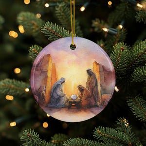Puede incluir: Adorno navideño redondo con una pintura a la acuarela de la escena de la Natividad. La imagen muestra tres figuras con túnicas reunidas alrededor de un bebé en un pesebre, con una luz brillante en el fondo. El adorno cuelga de un hilo dorado.