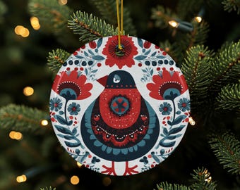 Könnte beinhalten: Runder Weihnachtsschmuck mit einem Vogeldesign im Folk-Art-Stil. Der Schmuck hat einen weißen Hintergrund mit roten Blumen, blauen Blättern und einem blauen und roten Vogel. Zum Aufhängen ist eine goldene Schnur befestigt.