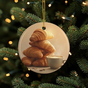 Peut inclure: Ornement de Noël rond représentant une pile de croissants dorés et une tasse de café. L'ornement est suspendu à un fil doré et se détache sur un fond doux et chaleureux. L'image évoque une scène de petit-déjeuner chaleureuse.