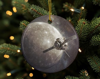 May include: Round Christmas ornament featuring a detailed image of a spacecraft in front of a planet and a starry background. The ornament is hanging from a gold string against a backdrop of a Christmas tree with lights.