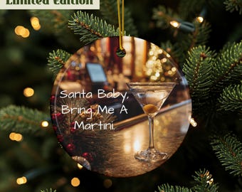 Può includere: Ornamento natalizio rotondo con l'immagine di un bicchiere da martini e la scritta "Santa Baby, Bring Me A Martini." L'ornamento ha un cordino dorato per appenderlo ed è circondato da rami di pino.