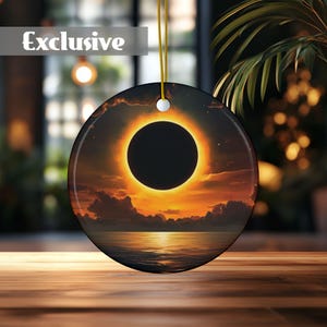 Op de afbeelding: Een ronde keramische ornament met een afbeelding van een zonsverduistering boven een watermassa. De verduistering is een ring van vuur met een donker centrum. De lucht is fel oranje en geel met wolken.
