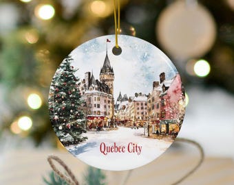 May include: A round ceramic ornament featuring a watercolor illustration of a snowy Quebec City streetscape. The scene includes a tall building with a pointed roof, a Christmas tree, and the text "Quebec City".