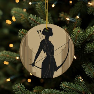 May include: Round Christmas ornament featuring a black silhouette of a woman archer with a bow and arrow, set against a cityscape backdrop. The ornament has a gold string for hanging and is set against a backdrop of a Christmas tree.