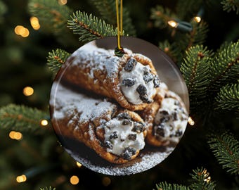 May include: Round Christmas ornament featuring a close-up of three cannoli pastries dusted with powdered sugar. The cannoli have a creamy white filling with dark chocolate chips. A gold string is attached for hanging.