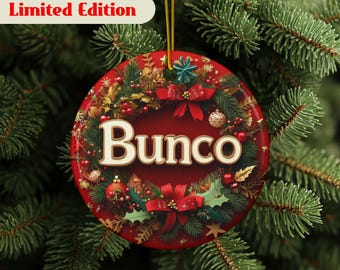 May include: A round, red Christmas ornament with the word "Bunco" in white script. The ornament is decorated with festive greenery, red bows, and gold accents. The text "Limited Edition" is displayed on a white banner.