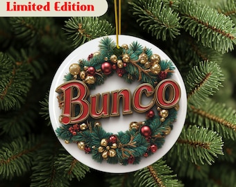 May include: A white ceramic Christmas ornament with the word "Bunco" in red, surrounded by a wreath of green pine needles, red berries, and gold accents. The ornament is hanging from a gold string, with the text "Limited Edition" at the top.