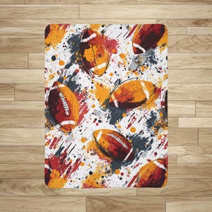 Puede incluir: Una manta rectangular blanca con un patrón repetido de balones de fútbol americano estilizados en marrón y naranja. El diseño incluye salpicaduras de rojo, naranja y gris, creando un efecto dinámico y artístico. Para los fanáticos del deporte.