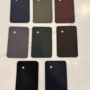 May include: Eight rectangular leather swatches arranged in three rows. The colors include navy blue, olive green, burgundy, muted purple, charcoal gray, dark brown, and two shades of black. Each swatch features a single circular hole at the top.