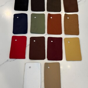 May include: A collection of fourteen rectangular leather swatches arranged in rows. The swatches feature a variety of colors including black, dark brown, olive green, rust, tan, navy blue, red, burgundy, yellow, white, and light beige.