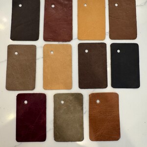 May include: Eleven rectangular leather swatches arranged in three rows. Each swatch features a single circular hole near the top edge. The colors include various shades of brown, tan, black, deep red, olive green, and a dark burgundy tone.