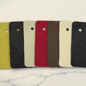 May include: A set of seven rectangular leather swatches arranged in a row, featuring a variety of colors including lime green, black, cream, red, brown, off-white, and dark navy blue. Each swatch has a single circular hole punched near the top.
