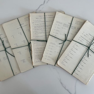 May include: A stack of vintage math worksheets, bound with green twine. The pages are aged with visible wear and tear, featuring printed equations and instructions. The worksheets are from a unit on whole numbers.