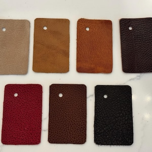 May include: Seven rectangular leather swatches arranged in two rows. The colors include beige, tan, light brown, dark brown, red, reddish-brown, and black. Each swatch features a single circular hole punched near the top center of the piece.