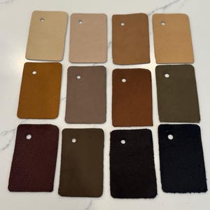 May include: A collection of twelve rectangular leather swatches arranged in three rows of four. Each swatch features a single circular hole near the top and displays a unique color, ranging from light beige and tan to brown, olive, burgundy, and black.