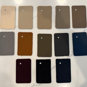 May include: A collection of twelve rectangular leather swatches arranged in three rows. The colors include cream, beige, tan, taupe, brown, gray, mustard, olive, charcoal, navy blue, deep burgundy, and black, each featuring a single punched hole.