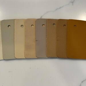 May include: A set of eight rectangular leather swatches arranged in a row, displaying a gradient of neutral tones ranging from off-white and light gray to beige, tan, and a deep golden brown. Each swatch features a single circular hole at the top.