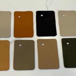 May include: Eight rectangular leather swatches arranged in two rows of four. The colors include light gray, mustard yellow, black, beige, tan, olive green, light brown, and dark forest green. Each swatch features a single circular hole punched near the top.