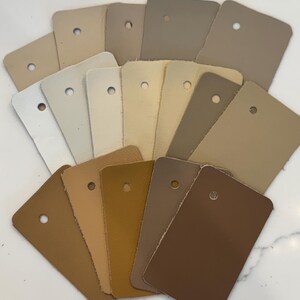 May include: A collection of rectangular leather swatches arranged in a fan shape. The swatches feature various shades of neutral colors, including cream, beige, tan, taupe, and brown. Each individual leather piece has a single circular hole punched near the top edge.