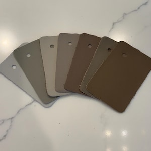 May include: A fan-shaped collection of seven rectangular leather swatches arranged in a gradient from light gray to dark brown. Each swatch features a single circular hole punched near the top edge, showcasing a smooth, matte-textured surface.