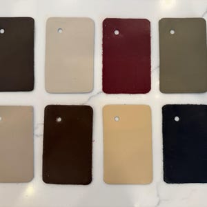 May include: Eight rectangular leather swatches arranged in two rows of four. Each swatch features a single circular hole near the top edge. The colors include dark brown, cream, deep burgundy, olive green, light tan, chocolate brown, beige, and black.