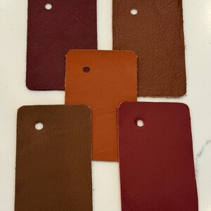 May include: Five rectangular leather swatches arranged in a cluster. The colors include deep burgundy, medium brown, bright orange, dark chocolate brown, and vibrant red. Each piece of leather features a single circular hole punched near the top.