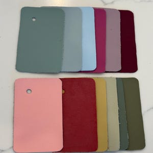 May include: A collection of rectangular leather swatches arranged in two rows. The top row features shades of sage green, light gray, sky blue, magenta, mauve, and deep burgundy. The bottom row includes light pink, red, mustard yellow, beige, olive green, and dark forest green.