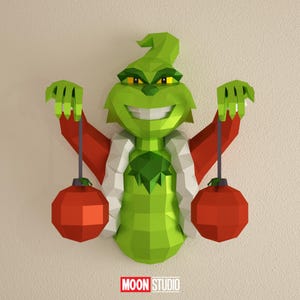 Puede incluir: Decoración de pared geométrica verde del Grinch, con brazos rojos y sosteniendo dos adornos rojos. El Grinch tiene un cuello blanco y una amplia sonrisa. El logotipo "MOON STUDIO" está en la parte inferior.