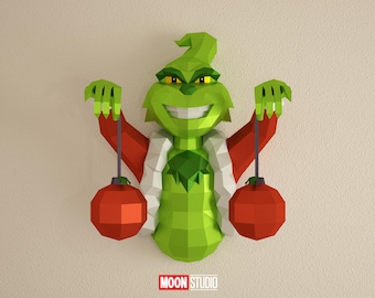 Puede incluir: Decoración de pared geométrica verde del Grinch, con brazos rojos y sosteniendo dos adornos rojos. El Grinch tiene un cuello blanco y una amplia sonrisa. El logotipo "MOON STUDIO" está en la parte inferior.