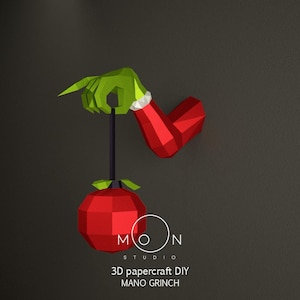 Puede incluir: Un modelo de papelcraft rojo y verde de la mano del Grinch sosteniendo un adorno rojo. El adorno está colgado de una cuerda. El texto "MOON STUDIO 3D papercraft DIY MANO GRINCH" está debajo del adorno.