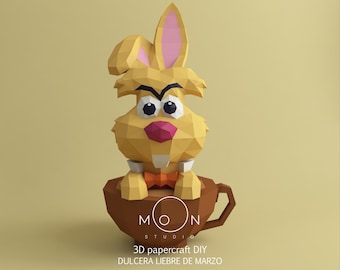 May include: A 3D papercraft DIY project of a yellow bunny rabbit wearing a bow tie and sitting in a brown teacup. The bunny has a grumpy expression and is holding a small white sign with the text "MOON STUDIOS". The text "3D papercraft DIY DULCERA LIEBRE DE MARZO" is below the image.