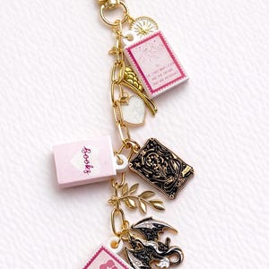 May include: Gold-coloured keychain with various charms. Charms include a black and gold tarot card, a dragon, and miniature pink books with text. The keychain also features a heart, a sunburst, and a sprig of leaves.