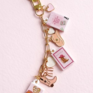 May include: A gold-coloured keychain featuring various charms, such as a pink cowboy boot and miniature books with text. The charms are linked by a gold chain, set against a pink backdrop.