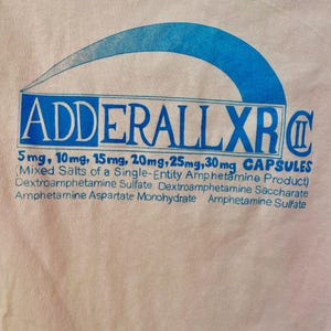 Può includere: Maglietta rosa chiaro con grafica blu. La grafica include il testo "ADDERALL XR" in un riquadro blu, con il testo "CAPSULES" sottostante. Il testo elenca anche i dosaggi: 5mg, 10mg, 15mg, 20mg, 25mg e 30mg.